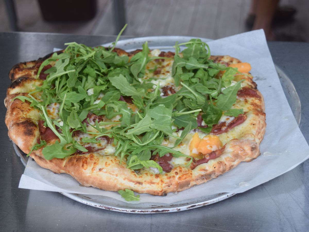 A pizza topped with arugula and ham on a white sheet of paper on a round metal tray. 