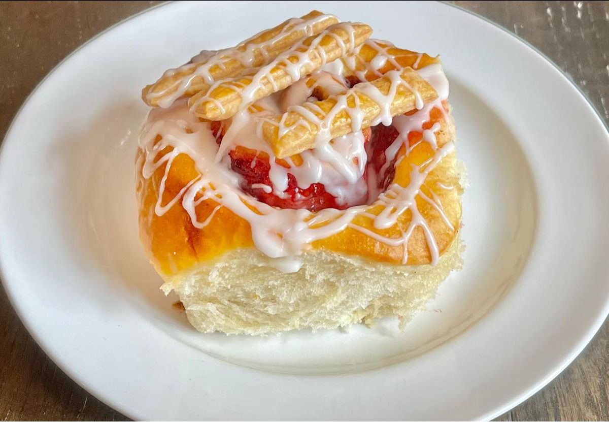 A kolache topped with lots of drizzled icing, jam, and toppings, on a white plate