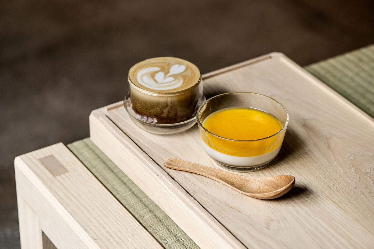A latte and side of orange juice colored drink on a wooden bench.