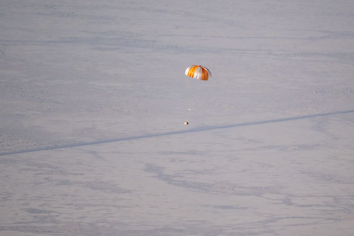 An orange and white parachute descends toward the desert ground