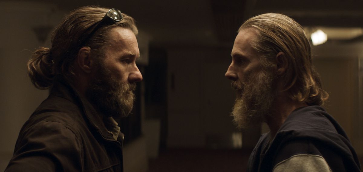(L-R) A bearded man (Joel Edgerton) with sunglasses on his head stands across and stares sternly at another bearded man (Sean Harris) in a hoodie.
