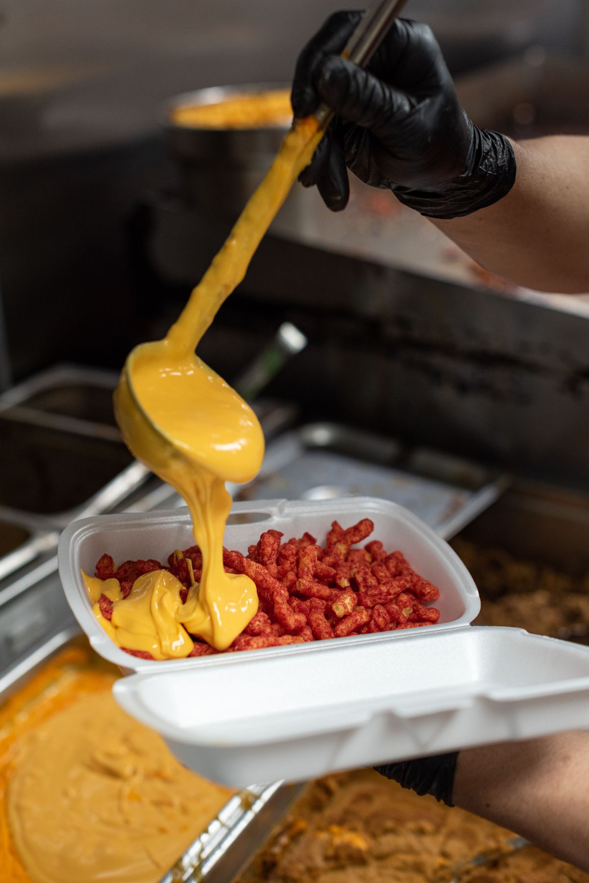 A ladle of cheese sauce over a styrofoam container of Hot Cheetos.