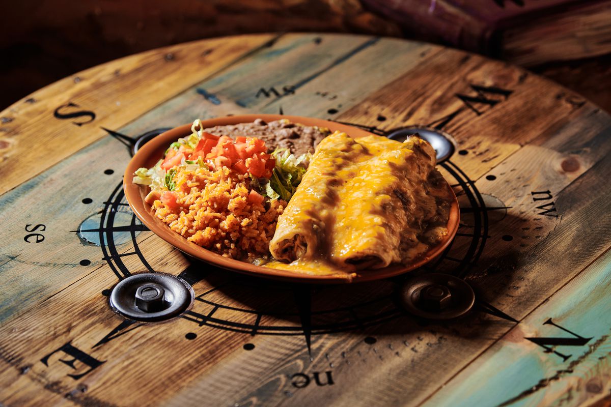 Green chile chicken echiladas served with orange rice with tomatoes and a side of beans on a tan plate, is presented atop a wooden table embossed with a compass decal.