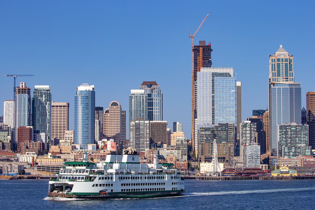 A lot of tall buildings, including ones under construction, in the background, and a passenger ferry on a harbor in the foreground. 