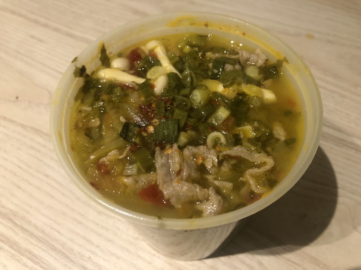 A quart container filled with a light yellow broth, shaved beef, white mushrooms, and green and red garnishes.