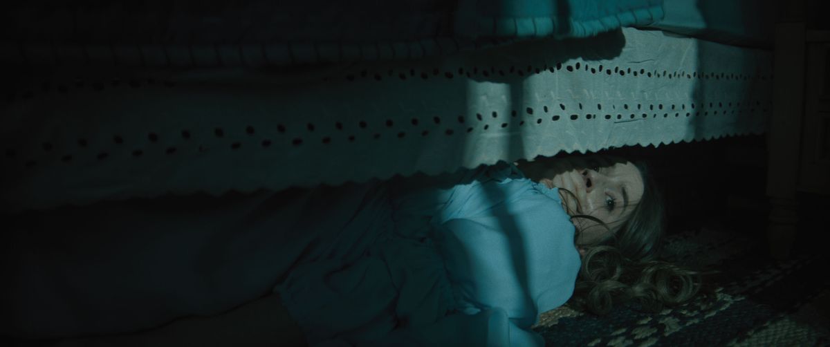 Brynn Adams (Kaitlyn Dever), a long-haired young woman in a blue nightgown, hides under her bed in the dark with her hand clapped over her mouth in the horror movie No One Will Save You