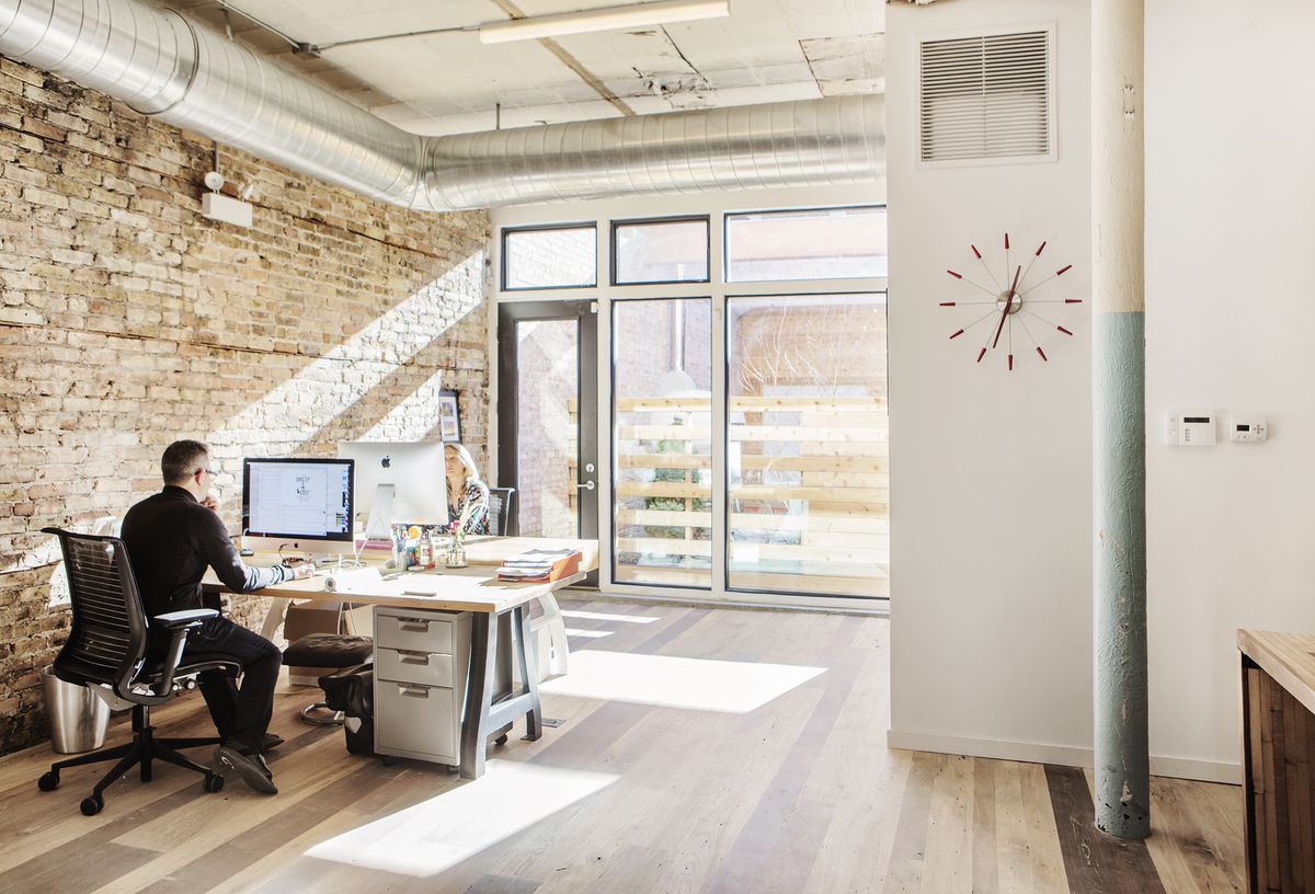An office area. One wall is exposed tan brick. There is a desk with a computer monitor. A man sits at the desk in an office chair. There are windows at the front of the space letting light in. On another white wall is a clock.