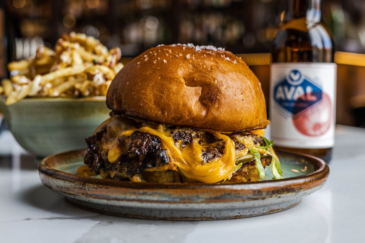 A cheeseburger in the foreground on a plate a bowl of fries to the left in the background and a brown bottle of beer to the right in the background from Bar Pigalle in Detroit, Michigan.