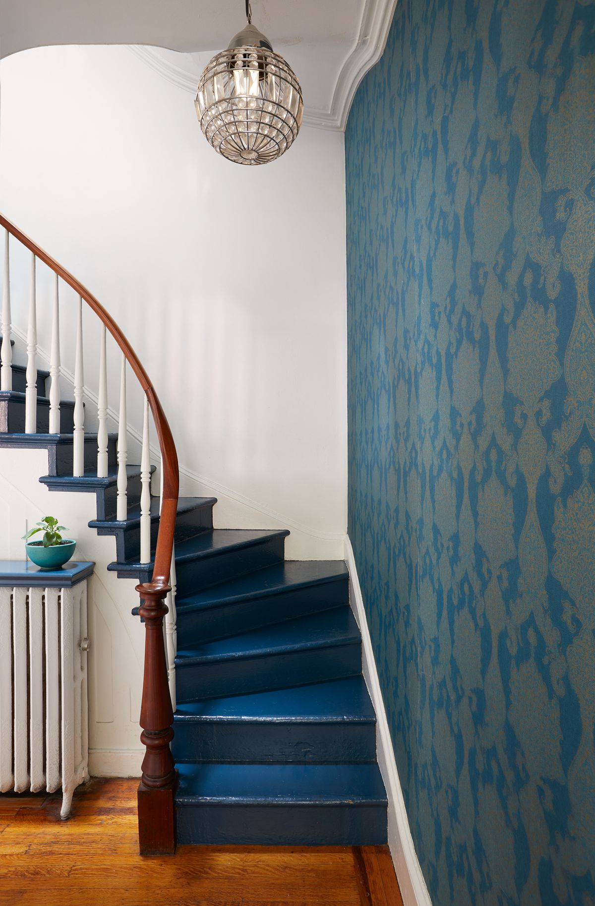 A staircase with blue stairs and a wooden bannister. There is patterned blue wallpaper on one wall. The other wall is painted white. There is a hanging light fixture.