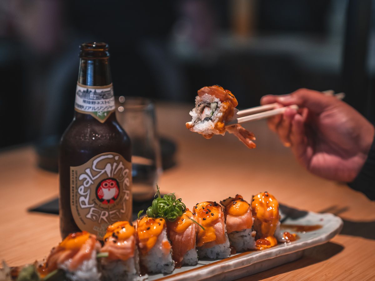 A sushi roll, glass brown beer bottle, a hand holding chopsticks holding a piece of sushi roll from TigerLily in Ferndale, Michigan.
