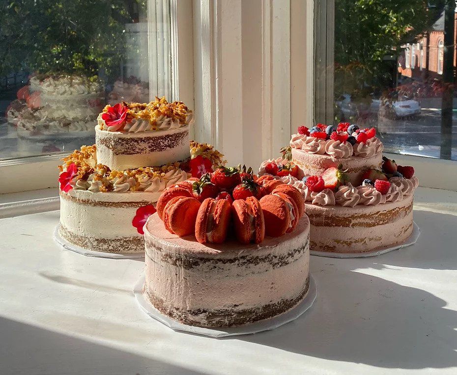 A variety of fruit-topped cakes, and one topped with full macarons, on a sill in a sunny window