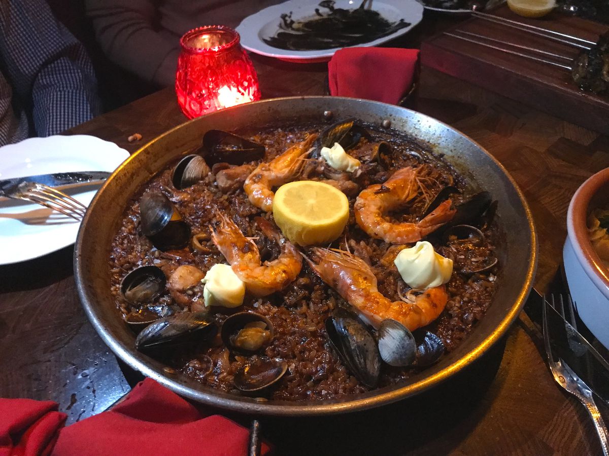 A round metal plan with paella, dark squid ink rice topped with seafood like shrimp, mussels, and clams with a half cut lemon on top.