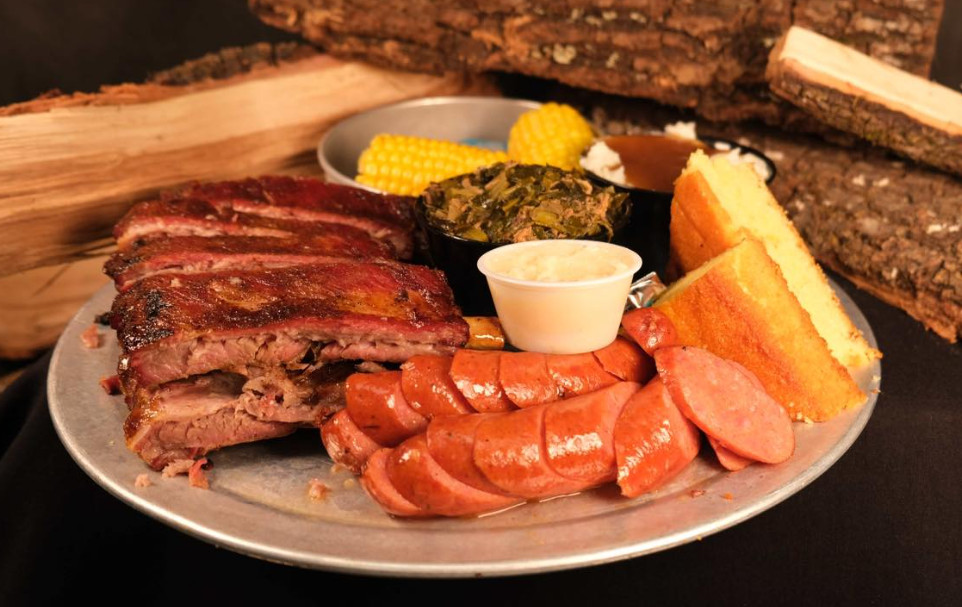 A platter of barbecue with ribs, sausage, cornbread, and corn, on a tin plate.
