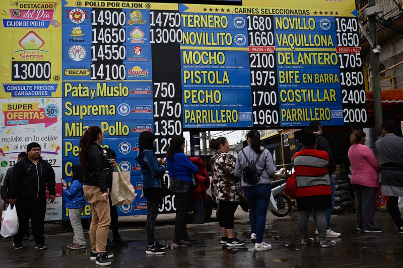 People stand in line on wet pavement in front of a large blue and yellow billboard listing different cuts of meat alongside their varying prices.