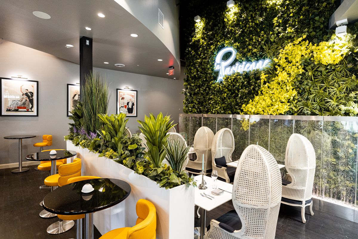 A dining room with yellow chairs, white wicker high-backed chairs, black and white topped tables, and a garden wall with yellow accents including a sign that reads: “Parisienne.”