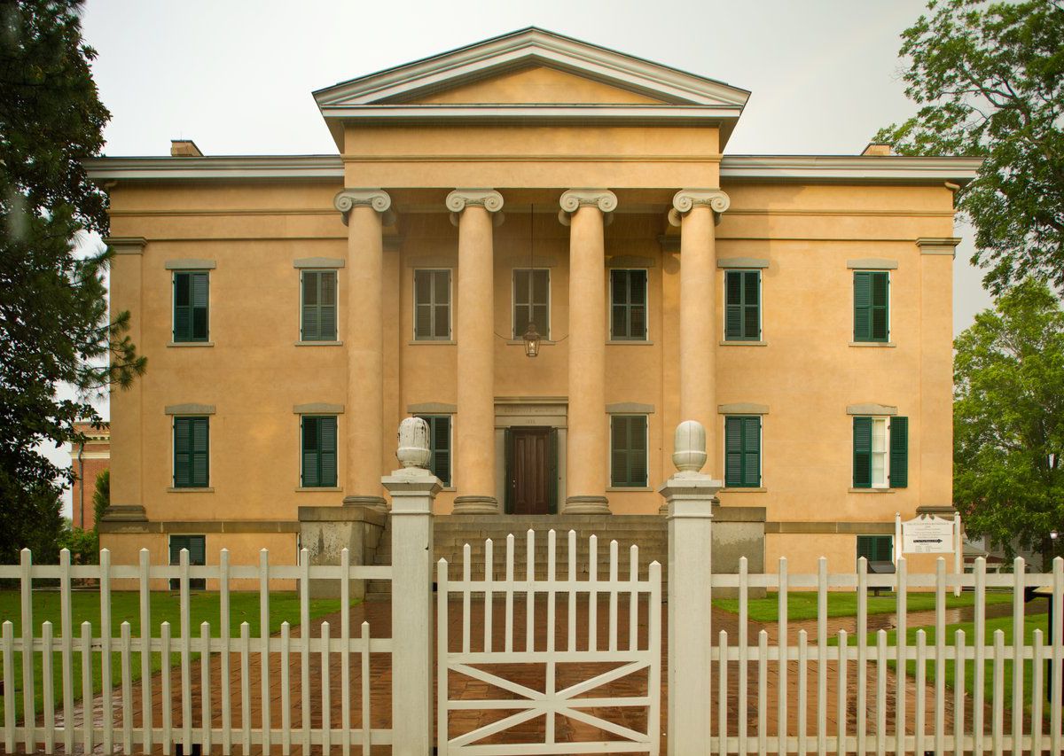 The exterior of Old Governors Mansion in Atlanta. The facade is yellow with columns in front. There is a fence and a lawn with trees in front of the house. 