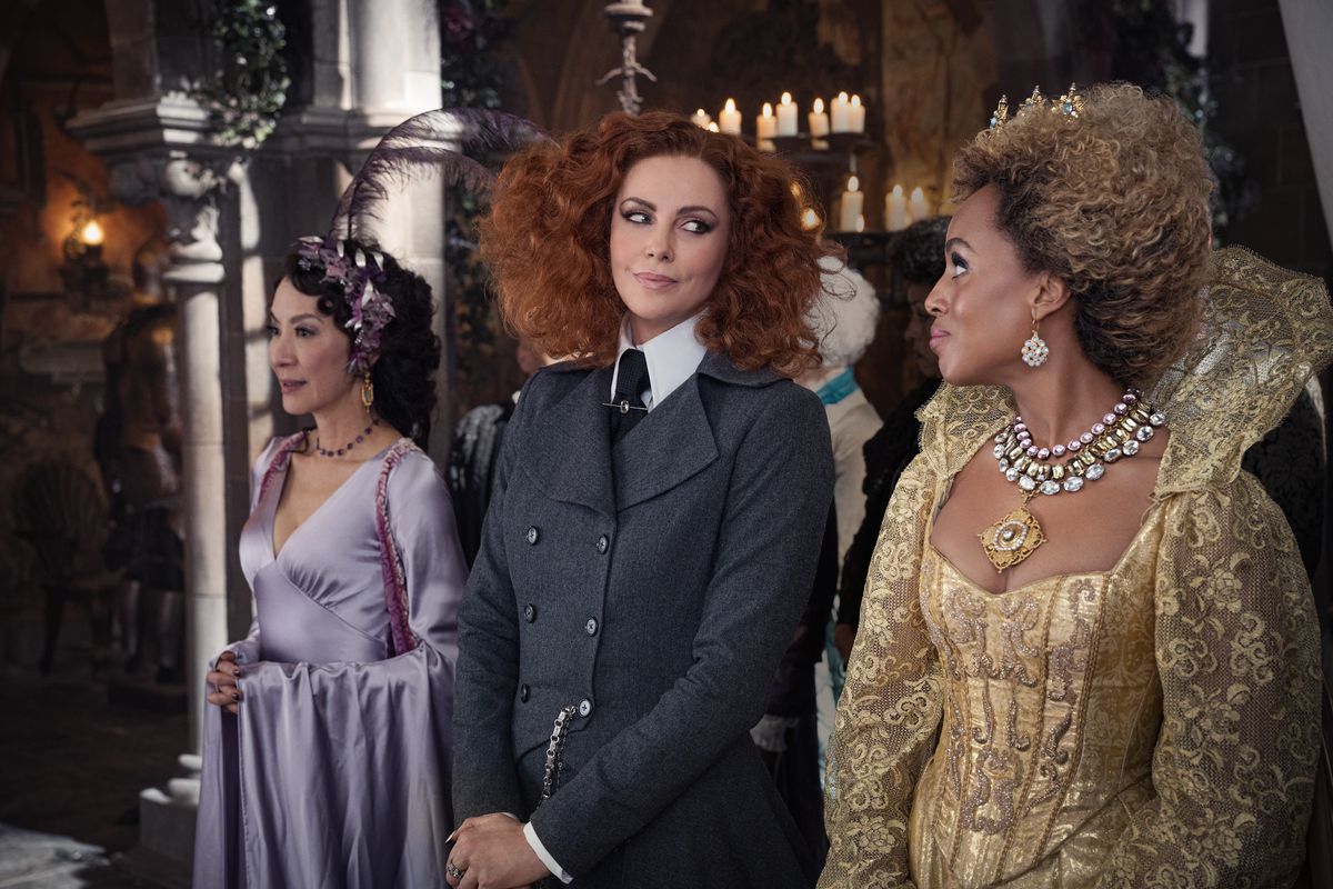 charlize theron in a dark suit and curly red hair casts a knowing glance at kerry washington, who wears a golden ballgown; behind them, michelle yeoh looks ahead in a purple dress in The School for Good and Evil