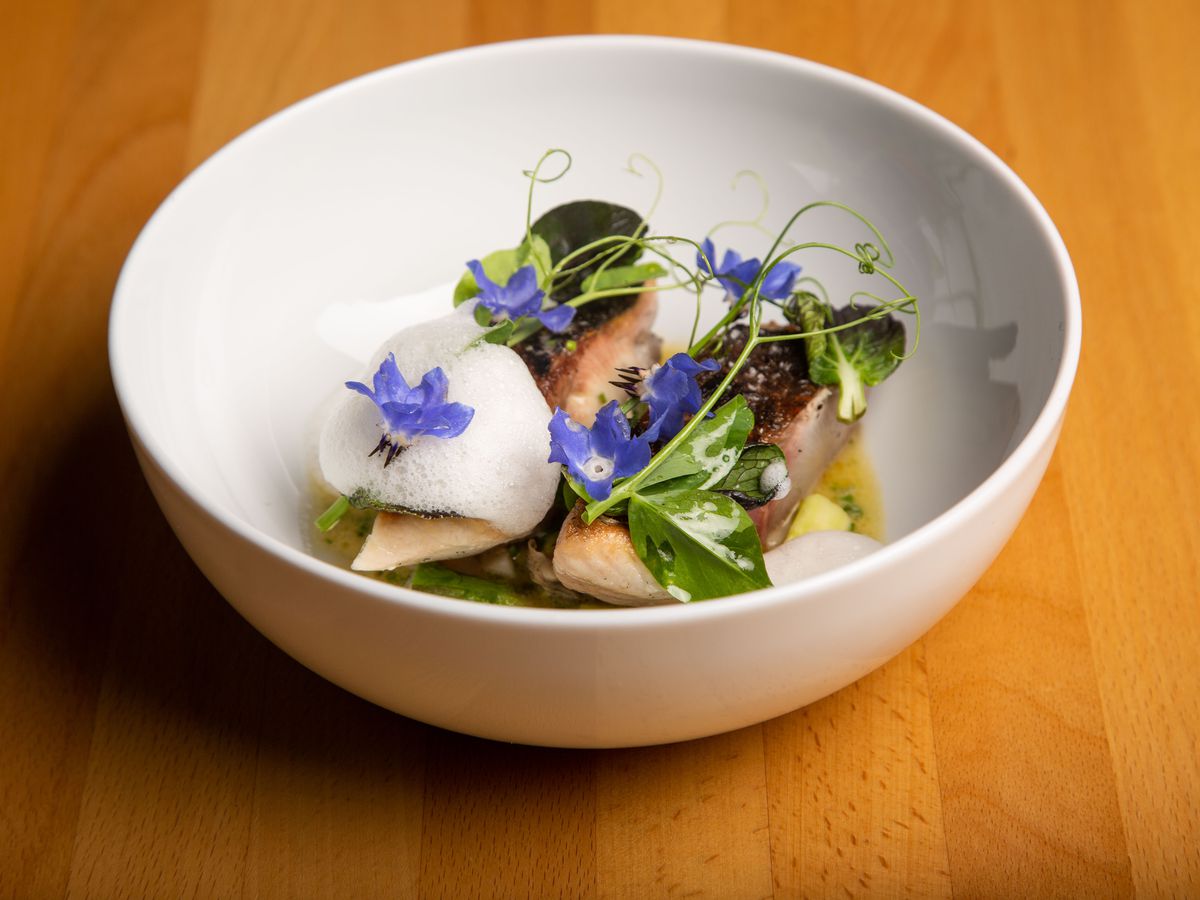 A deep bowl with several slices of fish beneath edible flowers and a puff ball of cream.