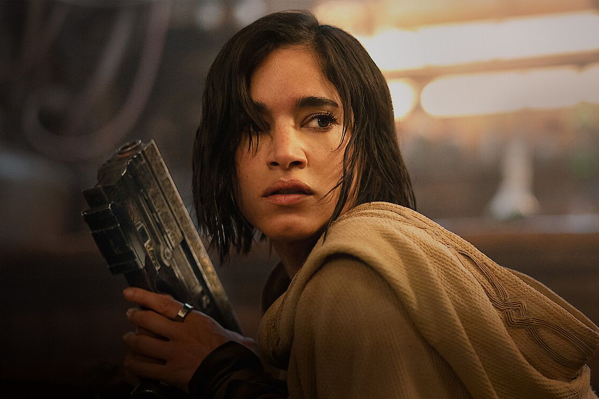 A close-up shot of a black-haired woman (Sofia Boutella) wearing a brown hooded shroud and holding a futuristic pistol in Rebel Moon.