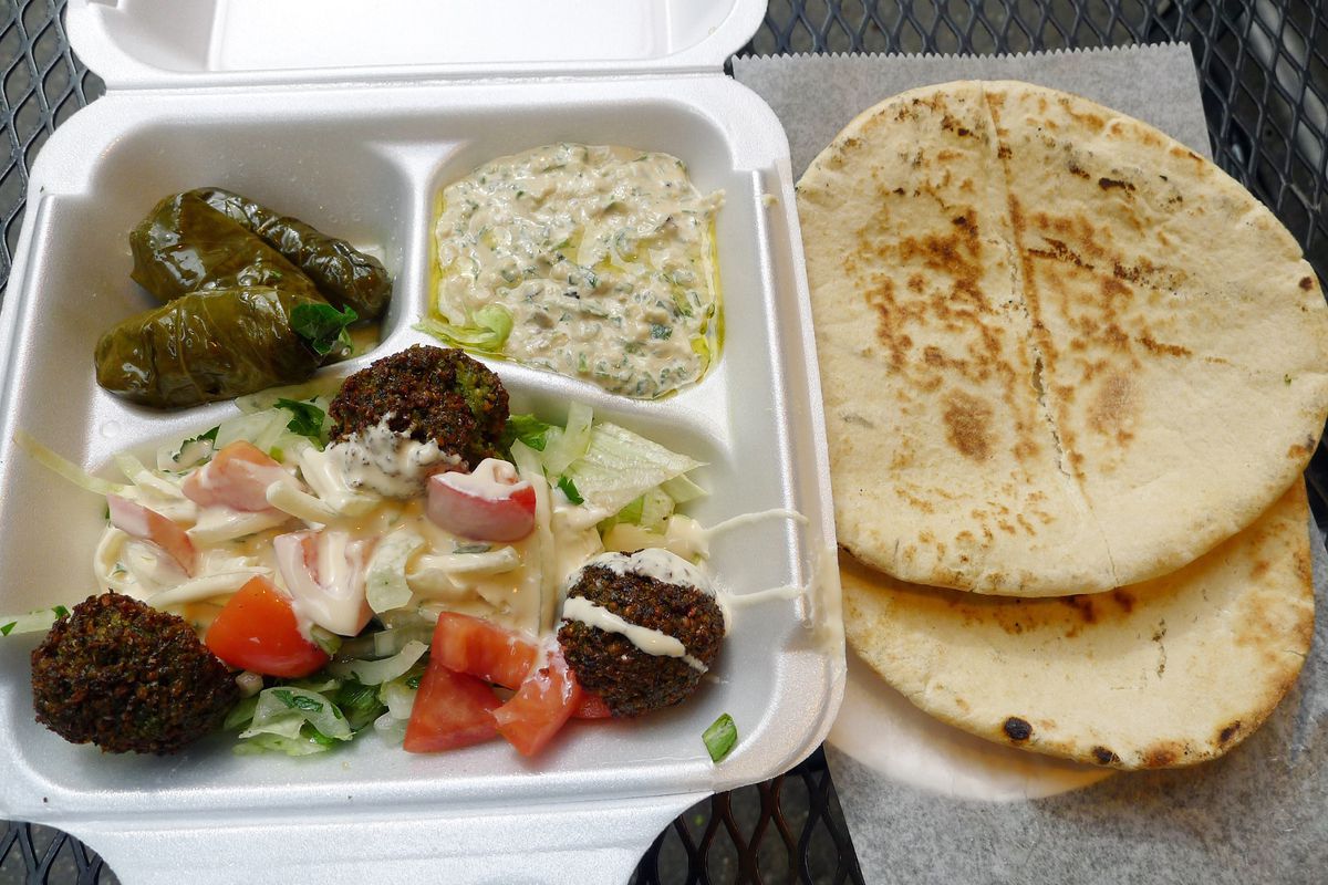 A styrofoam takeout container overflows with dolmas, falafel, and other dishes beside a stack of fluffy pita.