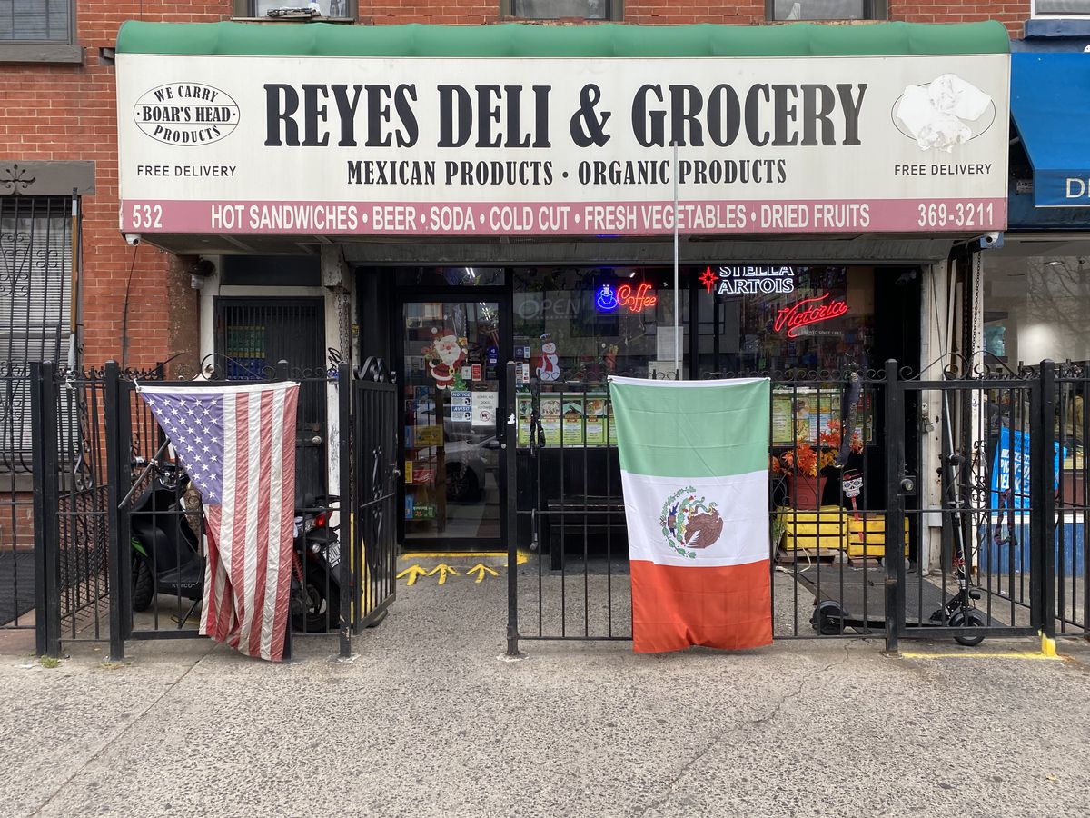 An exterior shot of a deli and grocery store with American and Mexican flags hanging on a front gate.