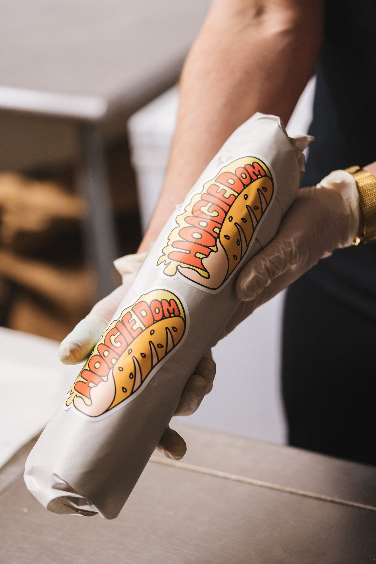 Two hands hold out a hoagie wrapped in paper with two stickers that say Hoagie Dom.