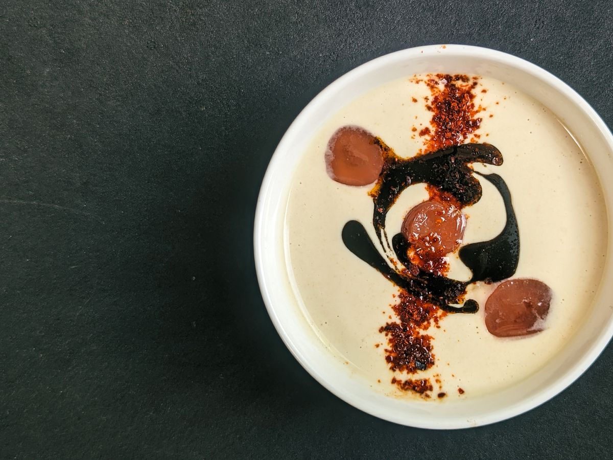 A bowl of blended almond gazpacho topped with grapes, black garlic oil, and a line of crushed pepper.
