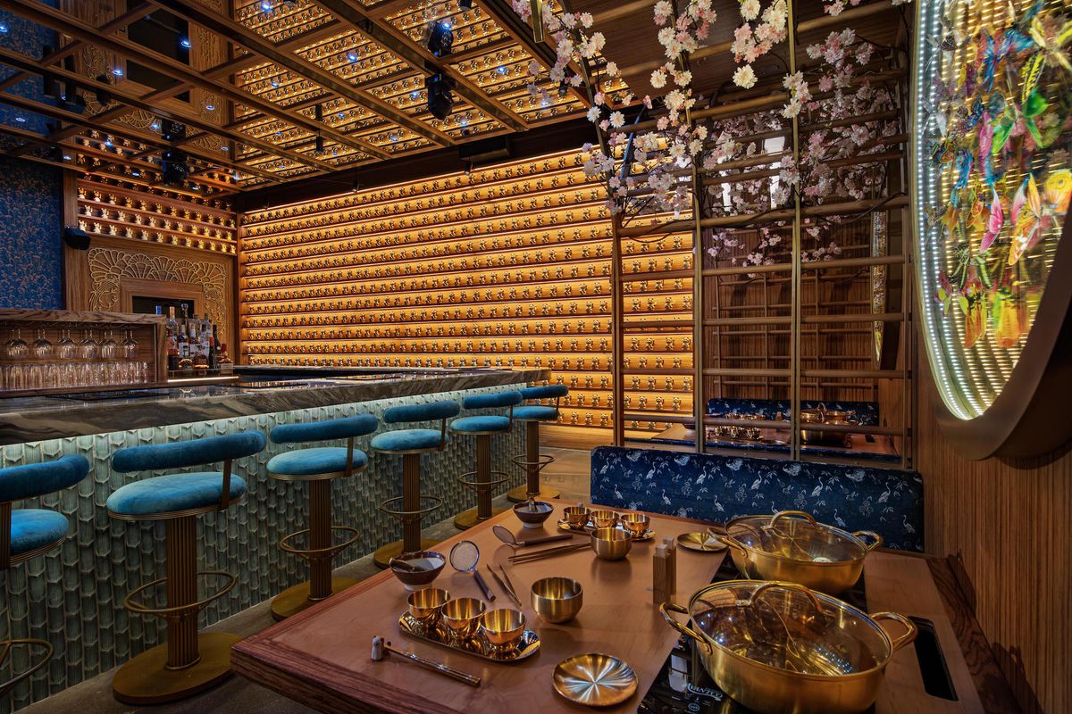 A tilted look at a wooden booth showing golden vessels for hot pot with teal bar seats in the background and a long golden wall with figurines at new LA restaurant Lucky Mizu.