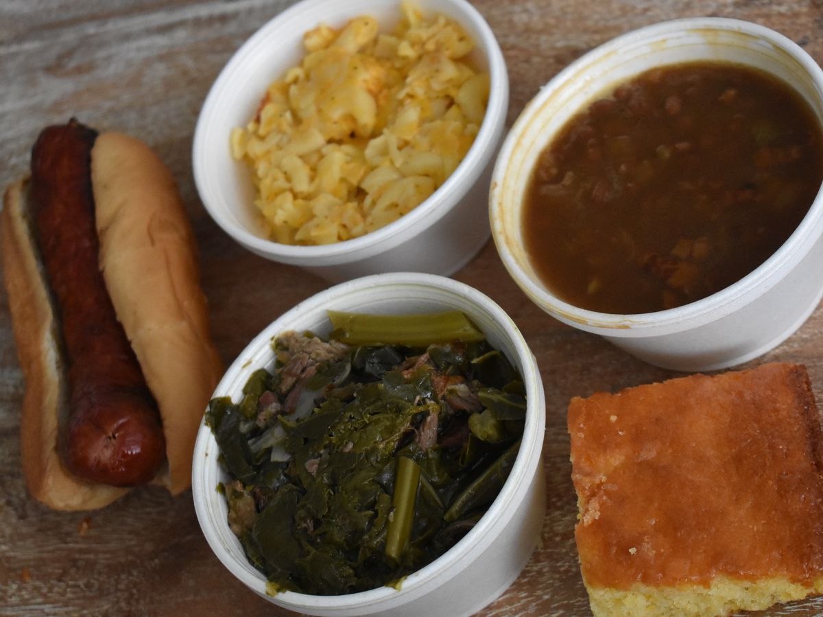 A sausage link in a hot dog bun is accompanied by three small white styrofoam bowls of collards, mac and cheese, and beans. There’s also a slice of cornbread.