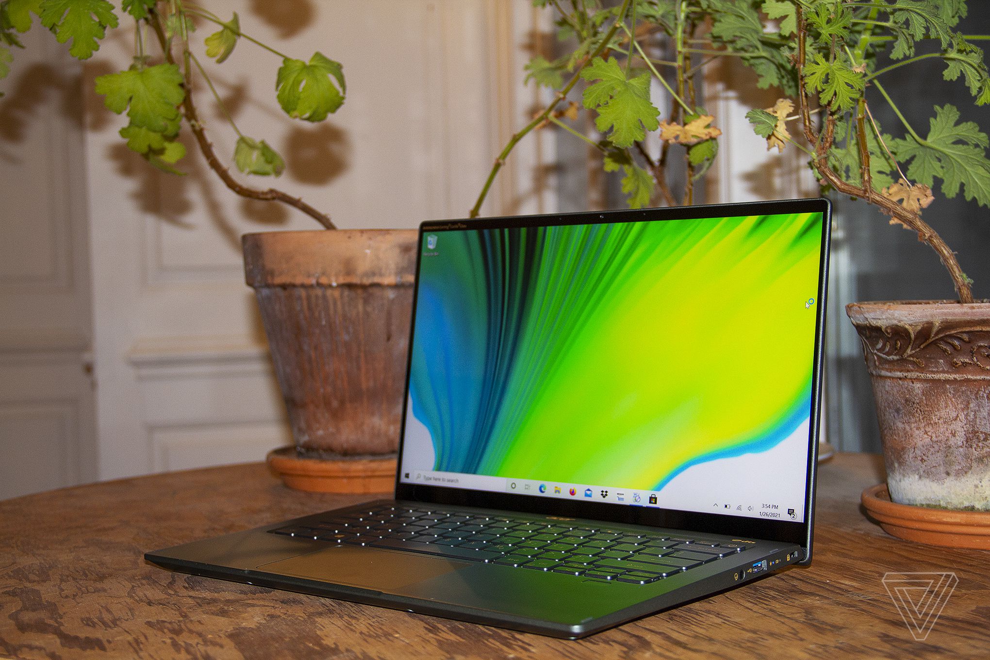 The Acer Swift 5 on a table in front of two house plants, open and angled to the left. The screen displays a green, blue, and white background.