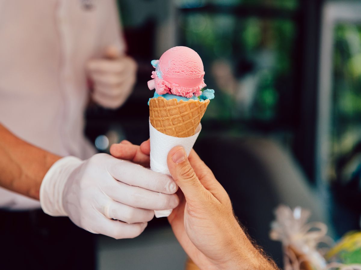 A gloved hand passes off an ice cream cone to an ungloved hand. It’s filled with pink and blue scoops of ice cream. 
