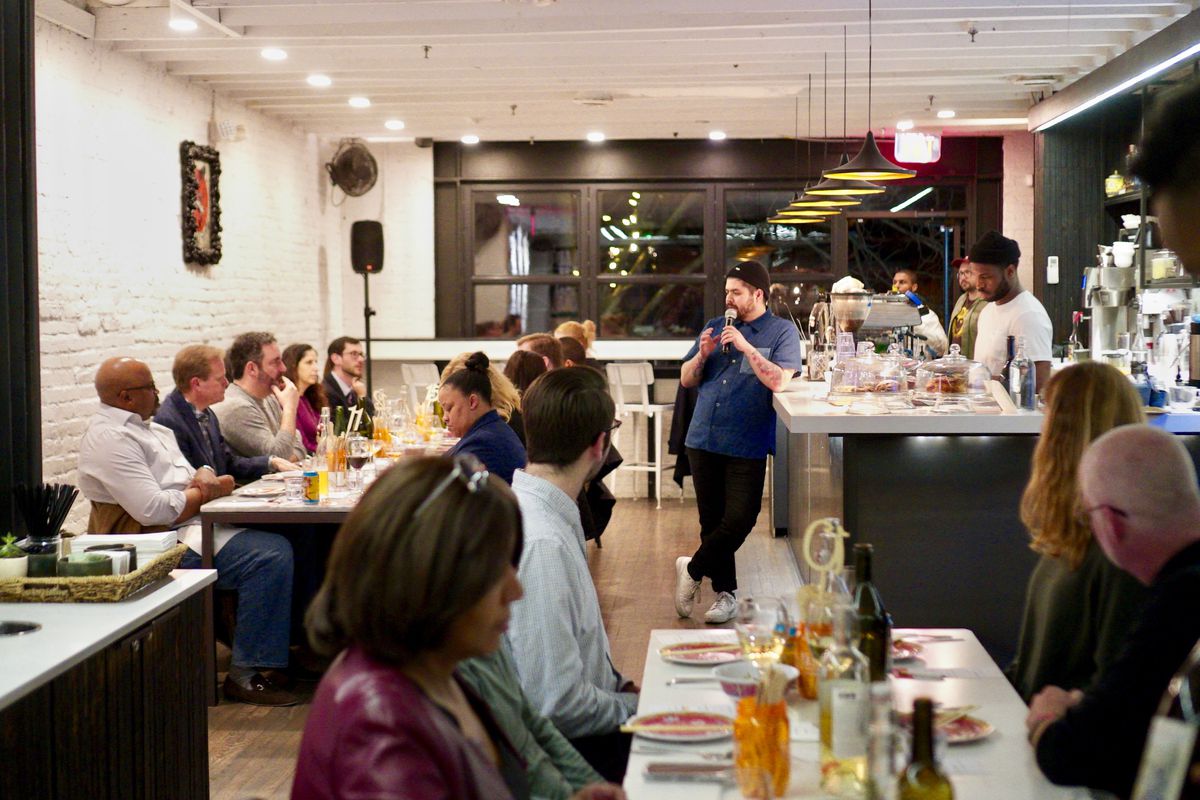 A chef speaks to diners seated at two communal tables.