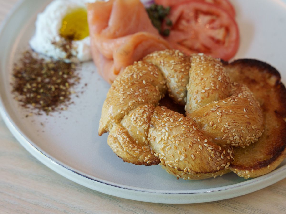 A braided bagel coated in sesame seeds on a plate with za’atar, lox, tomatoes, and labneh.