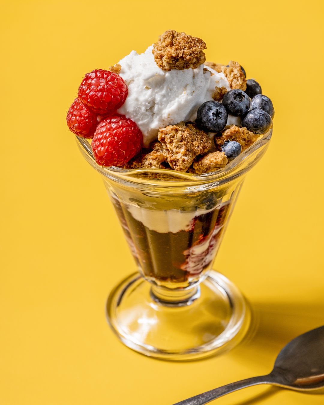 Coconut frozen yogurt in a clear parfait cup, topped with fruit, granola with a silver spoon next to the dish