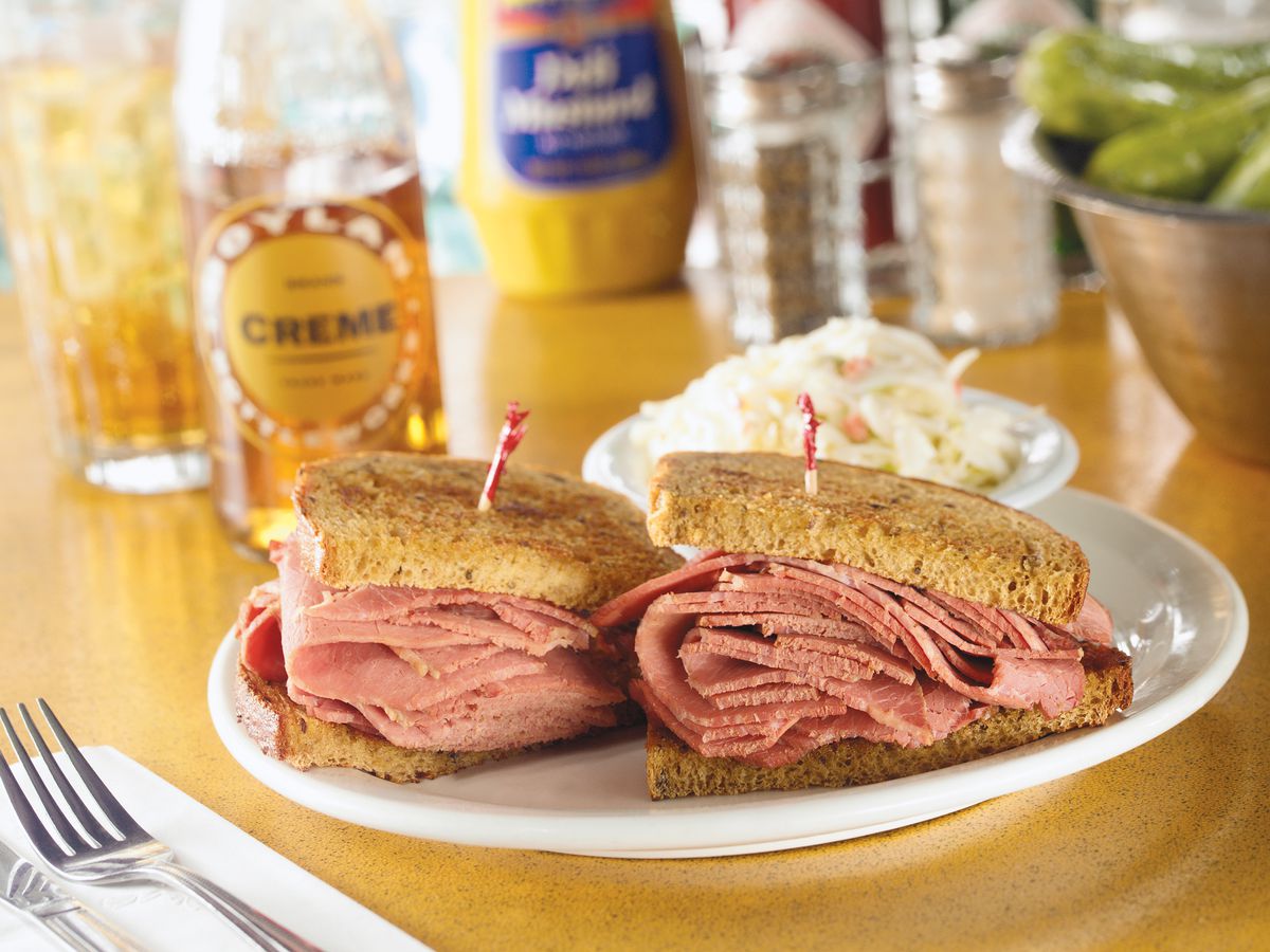 Two halves of a thickly stacked corned beef sandwich on a plate behind silverware on a napkin and in front of a dish of coleslaw and other slightly out-of-focus items like drinks, mustard, and pickles.