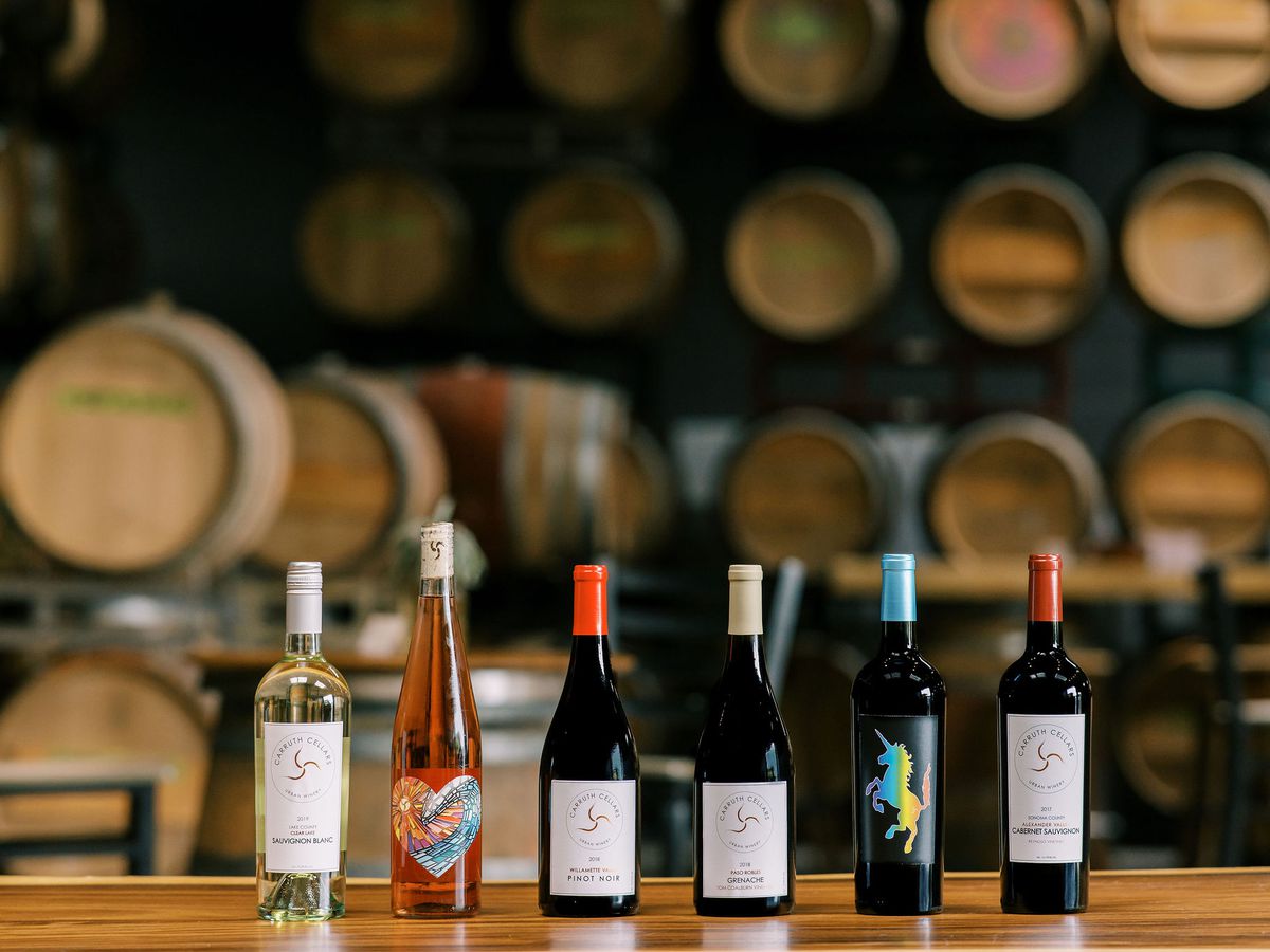 Six bottles of wine lined up against a backdrop of wooden barrels of wine. 