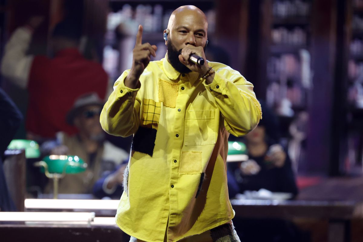Common performs onstage on November 8, 2023, in Inglewood, California, wearing an oversized, bright yellow button-up shirt and holding a microphone. 