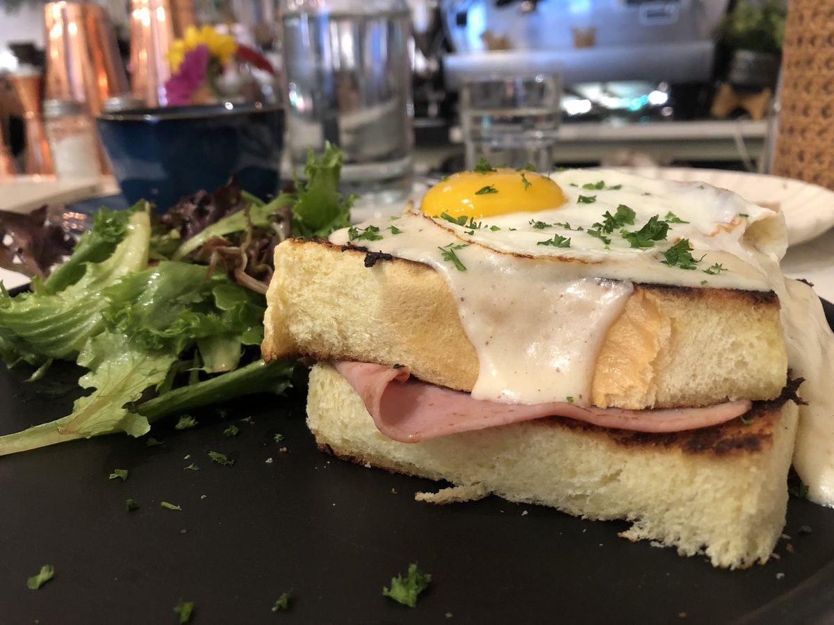Two slices of toasted bread with ham and cheese in the middle and a sunny side up egg on top. A small salad of lettuces is on the side of the plate.