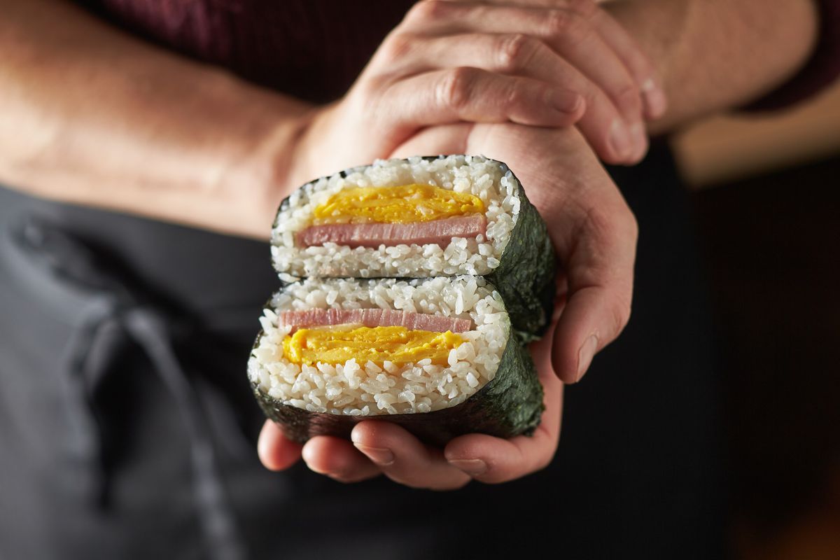 A hand holds a seaweed-wrapped ball of rice filled with ham and egg