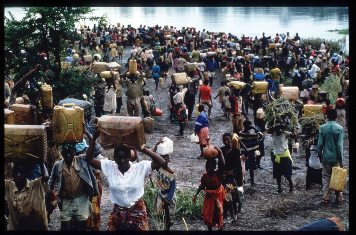 Crowds of people gather on a muddy riverbank, most holding belongings or balancing them on their heads.