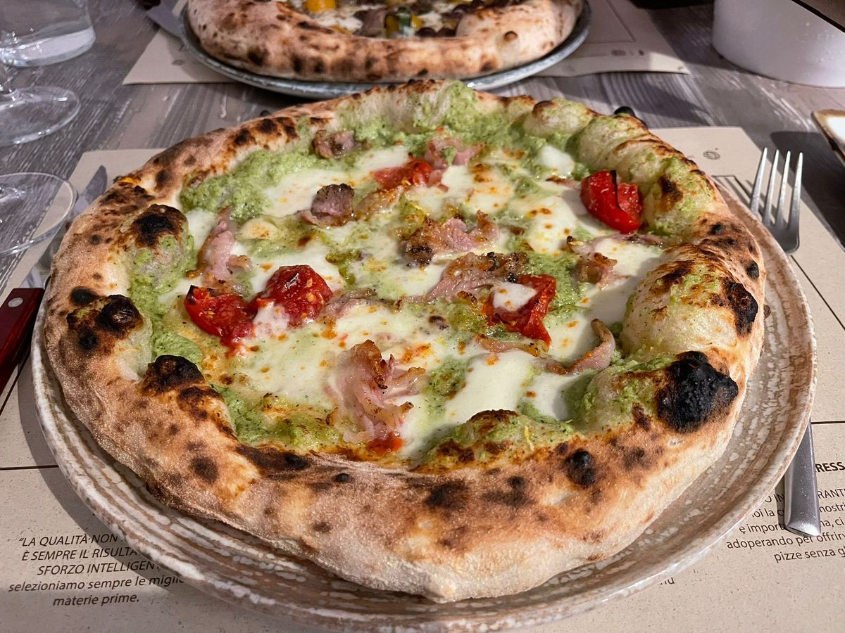 A whole pizza on a plate topped with green sauce, cheese, blistered tomatoes, and meat