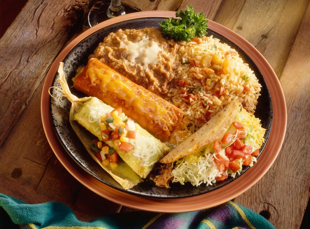 A plate of Mexican food, loaded clockwise with refried beans and rice, a taco, a tamale in a wrapper, and an enchilada in red sauce with cheese.