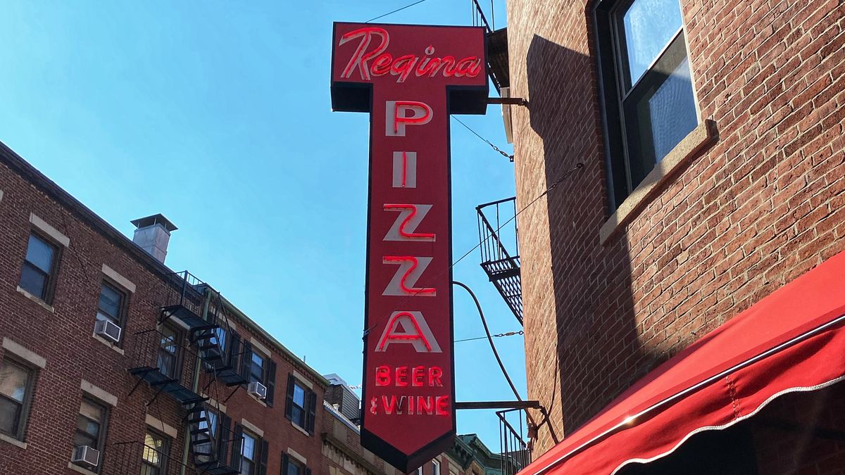 A sign for Regina Pizzeria hangs on the corner of a building with a red awning beneath the neon sign.