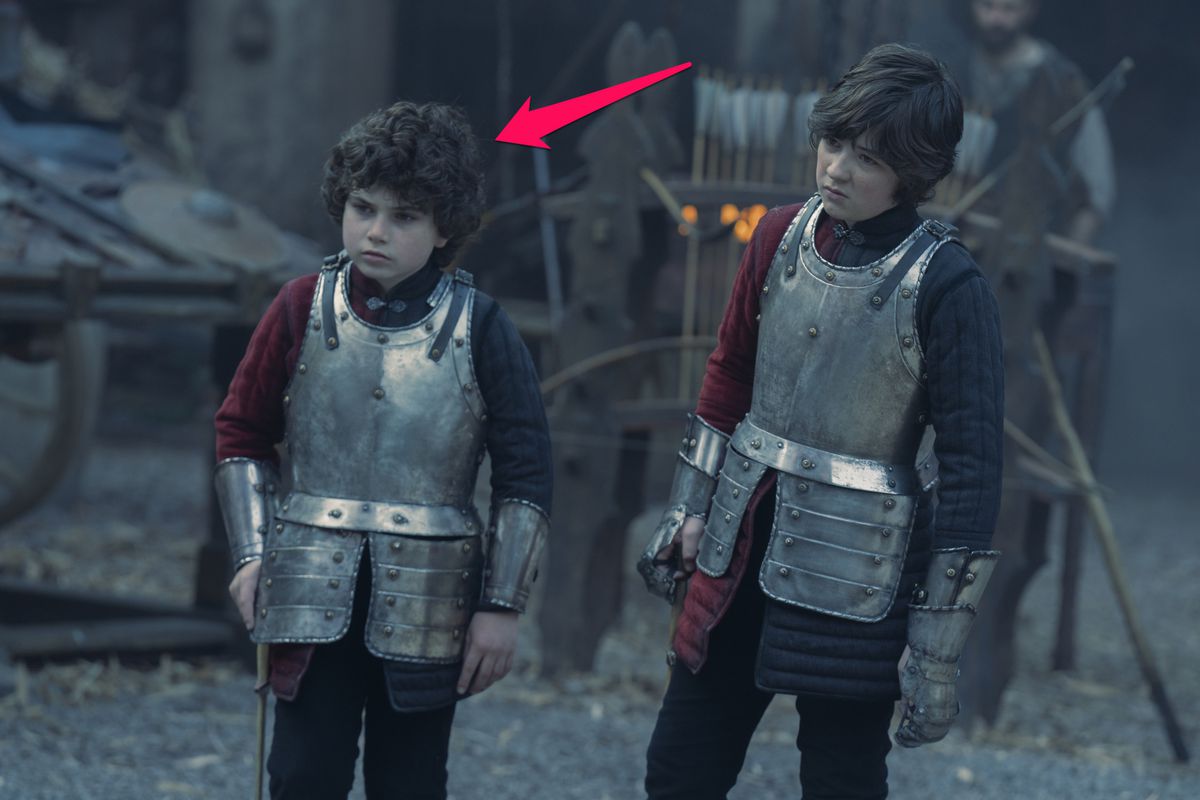 Harvey Sadler as Lucerys Velaryon from House of the Dragon.  A young boy with longer brown hair stands next to his brother, who has curly brown hair.  Both wear breastplates.  An arrow points to Lucerys, who has curly hair.