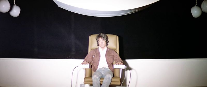 Warren Beatty as Lee Carter, sitting in a white chair staring at a bright screen.
