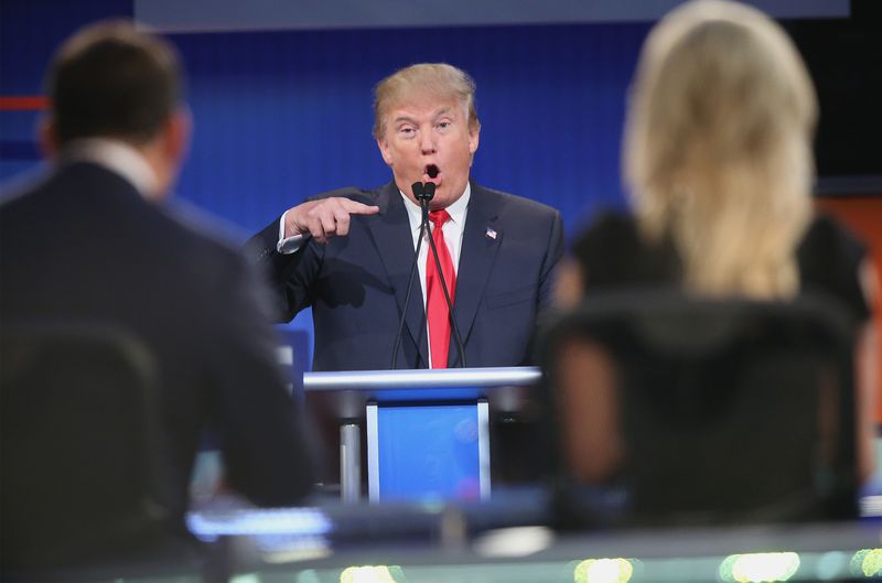 Konuşan ve parmağını yana doğru işaret eden Donald Trump, mikrofonla bir tartışma kürsüsünde duruyor.