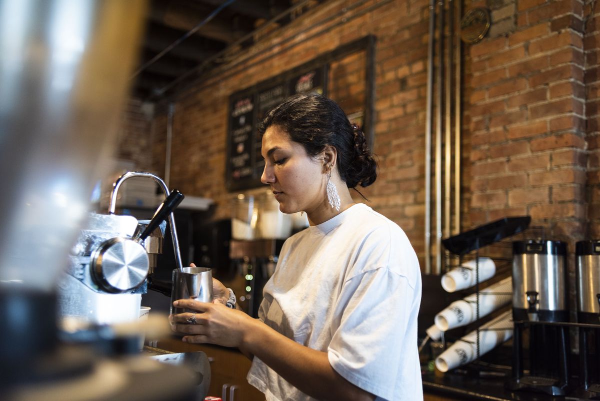 A woman with hair in a ponytail and a white t-shirt making a coffee drink with an espresso machine.