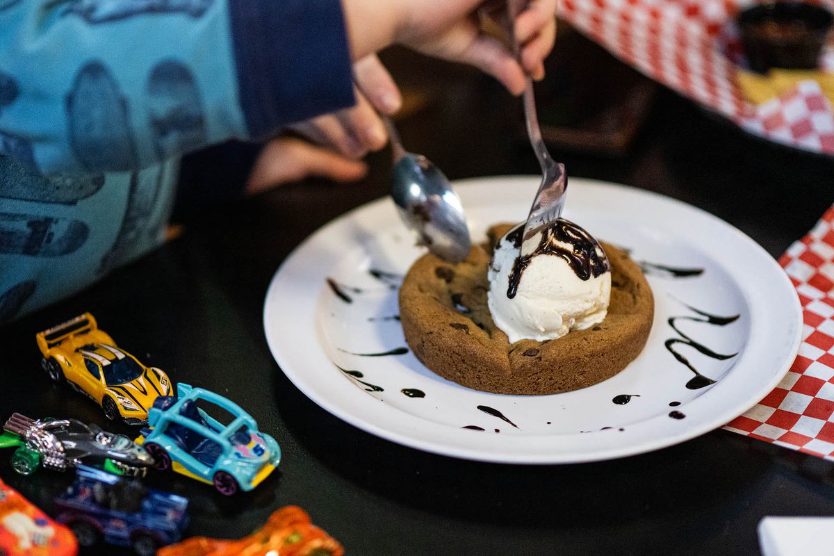 A child uses a fork and spoon to open a warm chocolate chip cookie underneath a scoop of ice cream on a white plate at a restaurant.