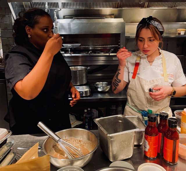 Two women in chefs aprons taste dishes with spoons in a kitchen.