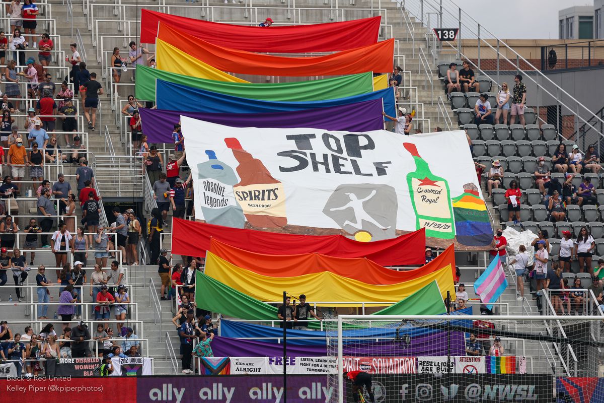 A tifo unfurled in the supporter’s section with pride colors at the top of bottom with a central image of glass bottles with one labeled ‘SPIRITS’ and the text ‘TOP SHELF’